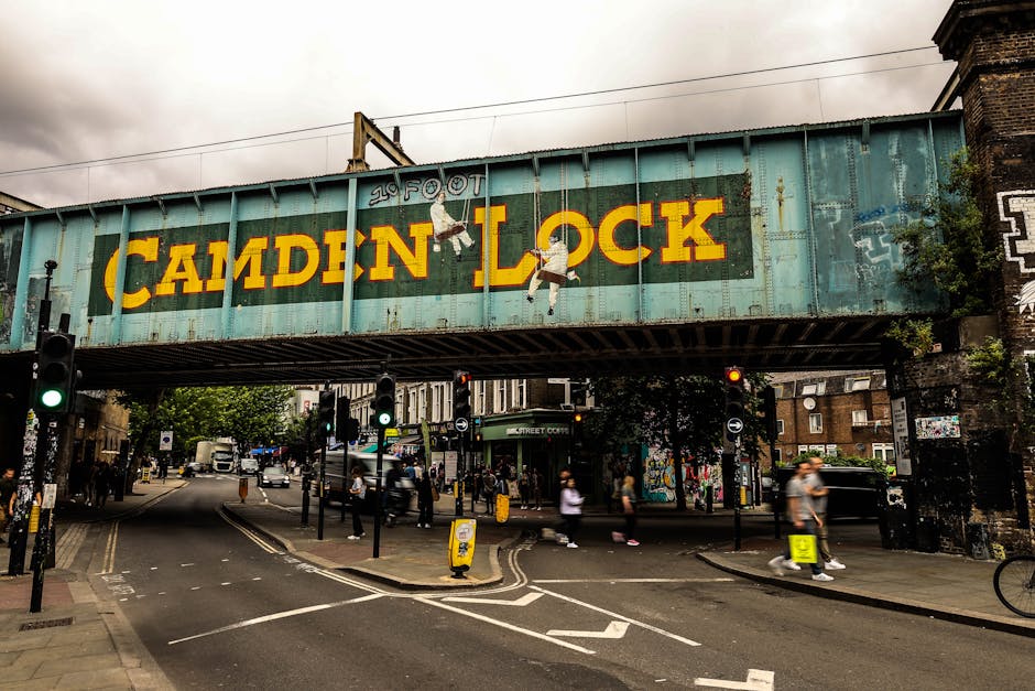 A large, weathered green metal railway bridge spans across a busy urban street, with yellow and black painted lettering reading 'Camden Lock' prominently displayed on the side. The bridge features two hanging graffiti-style figures, one holding a skateboard and the other appearing to be ascending, adding an artistic element to the structure. Below the bridge, the street is filled with pedestrians crossing at designated crosswalks, some walking briskly while others are standing at traffic signals. Multiple traffic lights are visible, with some showing a green signal, and a small yellow street sign is positioned on the pavement. To the right, part of an old brick building with graffiti accents and some greenery growing around its edge can be seen. The scene is captured in overcast weather, creating a diffuse light that emphasizes the urban atmosphere. The setting suggests an active area where independent waste collection or rubbish removal services, such as those offered by House Clearance Swiss Cottage, may facilitate debris clearance in a busy city context with a focus on private waste handling outside of municipal collection.