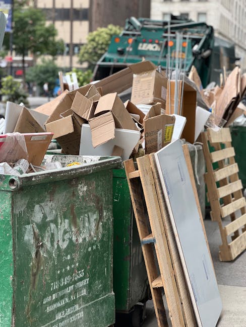 A large green wheelie bin filled with various types of waste, including cardboard boxes, some flattened and others partially assembled, along with some packaging materials. The boxes are made of brown corrugated cardboard, with a few white and patterned cardboard pieces visible among the pile. The waste spills over the top of the bin, which is situated on a city street in an urban environment, with a background showing a street scene, blurred trees, and part of a waste collection vehicle. To the right of the bin, there is a wooden pallet leaning against it, and additional cardboard sheets are stacked nearby. The scene suggests a private or independent rubbish collection, with clear evidence of waste that requires removal, typical of a house clearance or renovation cleanup. The overall atmosphere is neutral, with natural daylight illuminating the scene, highlighting the textures of the cardboard, the weathered surface of the bin, and the surrounding urban elements.