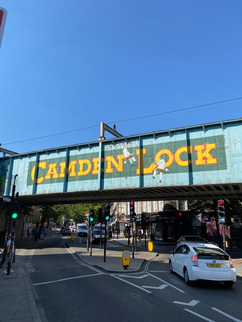 A large, weathered green metal railway bridge spans across a busy urban street, with yellow and black painted lettering reading 'Camden Lock' prominently displayed on the side. The bridge features two hanging graffiti-style figures, one holding a skateboard and the other appearing to be ascending, adding an artistic element to the structure. Below the bridge, the street is filled with pedestrians crossing at designated crosswalks, some walking briskly while others are standing at traffic signals. Multiple traffic lights are visible, with some showing a green signal, and a small yellow street sign is positioned on the pavement. To the right, part of an old brick building with graffiti accents and some greenery growing around its edge can be seen. The scene is captured in overcast weather, creating a diffuse light that emphasizes the urban atmosphere. The setting suggests an active area where independent waste collection or rubbish removal services, such as those offered by House Clearance Swiss Cottage, may facilitate debris clearance in a busy city context with a focus on private waste handling outside of municipal collection.