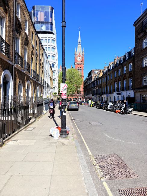 A city street scene in daylight with a wide concrete pavement on the left side, where a person is seen walking past a large white plastic bag of rubbish placed on the sidewalk. The pavement is lined with traditional brick terrace houses featuring black wrought iron railings and arched window and door openings. A tall, modern building with reflective glass windows stands behind the row of terrace houses on the left. In the background, the spire and clock tower of a historic red brick church or civic building is visible, rising prominently against a clear blue sky. Several vehicles, including cars and motorbikes, are parked along the street and some are in motion. A black street lamp pole is centrally positioned, with various road signs attached to it, including a pedestrian warning sign. The overall scene reflects urban waste management with the visible rubbish bag indicating an area where private or independent waste collection services, such as those offered by House Clearance Swiss Cottage, may handle rubbish removal outside local authority schedules in a typical London setting.