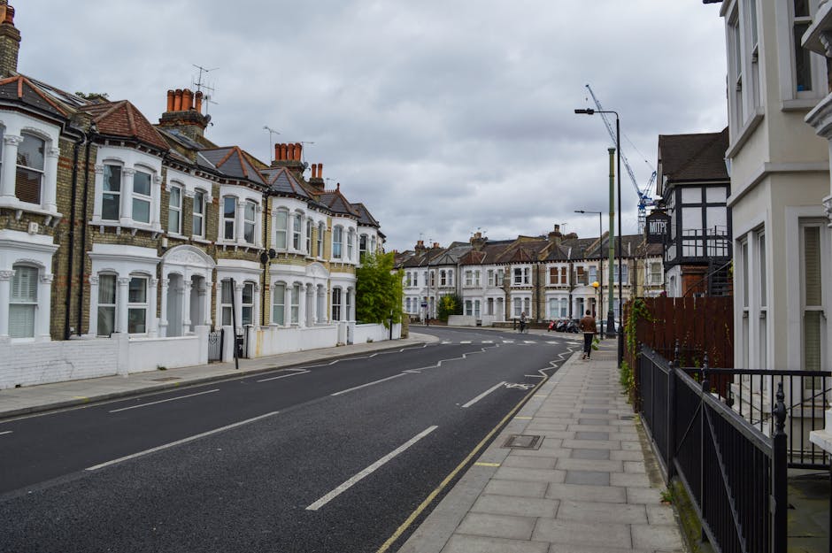 A residential street scene in a neighborhood with Victorian-style terraced houses featuring ornate white facades, large bay windows, and red-brick chimneys, set against an overcast sky. The pavement runs alongside row of well-maintained houses on the right, separated by black metal railings. In the foreground, a person dressed in dark clothing is walking on the sidewalk. The street is relatively quiet, with a few parked cars visible further along, and a construction crane is seen in the distance, indicating ongoing building work. The overall atmosphere is calm and typical of a suburban area in the UK, with neutral tones and no visible rubbish or waste, though the setting aligns with professional rubbish clearance and waste management services that handle local residential cleanouts and private disposal needs.