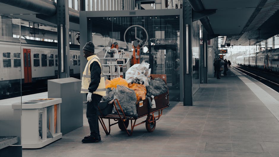A person wearing a dark jacket, grey beanie, and high-visibility safety vest stands beside a red handcart loaded with various garbage bags and discarded items on a modern train station platform. The bags appear to be made of plastic and fabric in shades of black, grey, and orange, with some containing recyclable materials or waste awaiting collection. Behind the person, there's a glass-enclosed structure with mechanical equipment and rail infrastructure, including train tracks and platform edge tactile paving on the right. The background shows an active train station environment with a stationary train on the left side and a few distant figures walking along the platform. The scene is lit with natural daylight, emphasizing the industrial setting and the presence of waste that may be targeted for removal or designated for private disposal services, aligning with the context of rubbish removal and on-site clearance managed by companies like House Clearance Swiss Cottage.