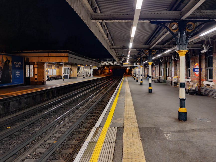 The image depicts a quiet underground train station platform at night, illuminated by overhead fluorescent lighting. The platform runs parallel to multiple railway tracks with visible wooden sleepers and metal rails extending into the distance. On the right side, there is a row of black and yellow painted bollards and columns supporting the station's roof structure, which features a corrugated metal ceiling with exposed beams. Behind the columns, brick walls and large windows are visible, some showing signs and posters, including the classic London Underground roundel sign. The left side of the platform includes a small brick building with windows, possibly a ticket office or waiting area, with a person seated on a bench nearby. The ground surface is composed of concrete with tactile paving strips near the platform edge, also marked with a yellow line for safety. The scene suggests an environment suited for both scheduled and alternative arrangements for train station access, with the presence of tailored infrastructure for passenger safety and guidance, fitting into the broader context of independent transit options and private platform management.
